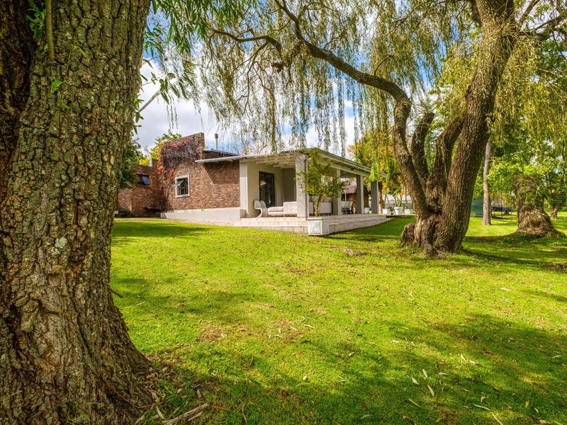 A view of the rear exterior of the house, featuring a covered patio area, brick facade, and expansive green garden with mature trees.