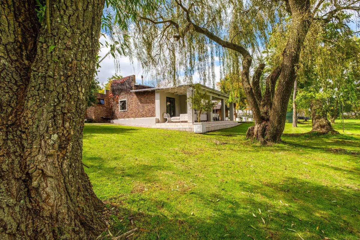 A view of the rear exterior of the house, featuring a covered patio area, brick facade, and expansive green garden with mature trees.