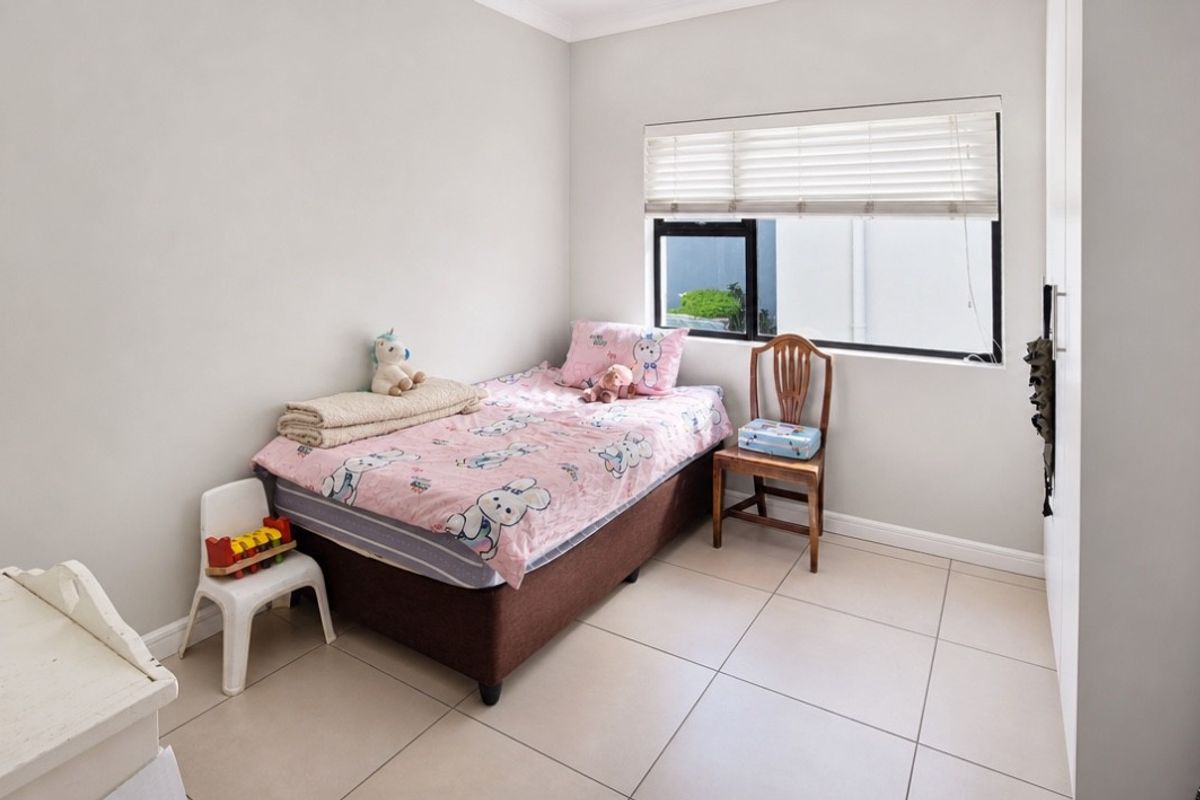 A bright bedroom featuring neutral tiled flooring, a built-in wardrobe, and a window fitted with venetian blinds.