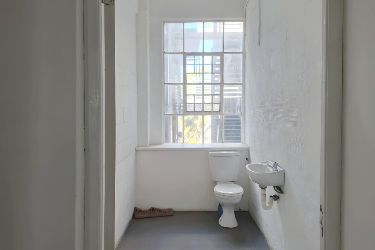 A compact office restroom featuring a toilet, wall-mounted basin, and large industrial-style window providing natural light.