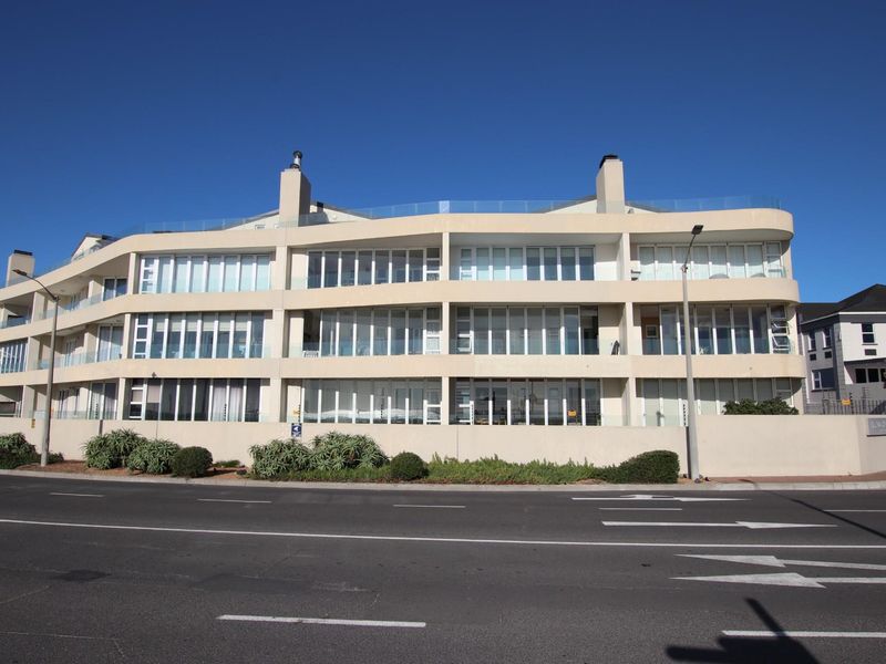 A clear street-level view of a modern multi-story apartment building featuring large glass windows, balconies with glass balustrades, and chimneys.
