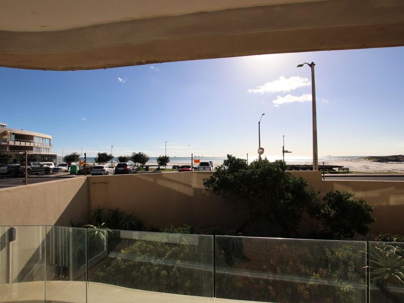 A view from a balcony overlooking a coastal road, parking area, and the ocean beyond.