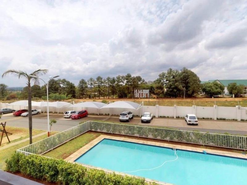Elevated view of the communal swimming pool area, featuring a fenced pool, landscaped garden, and covered parking bays within the apartment complex.