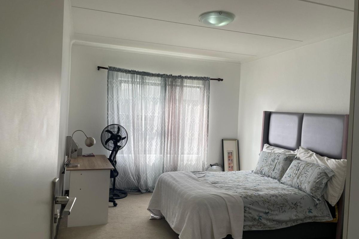 A well-lit bedroom featuring neutral carpeting, a large window with sheer curtains, and a central ceiling light fixture.