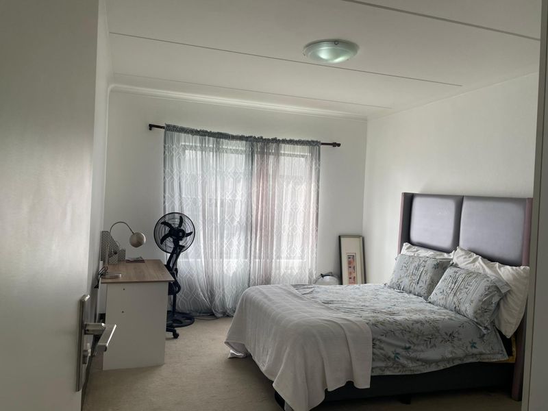 A well-lit bedroom featuring neutral carpeting, a large window with sheer curtains, and a central ceiling light fixture.