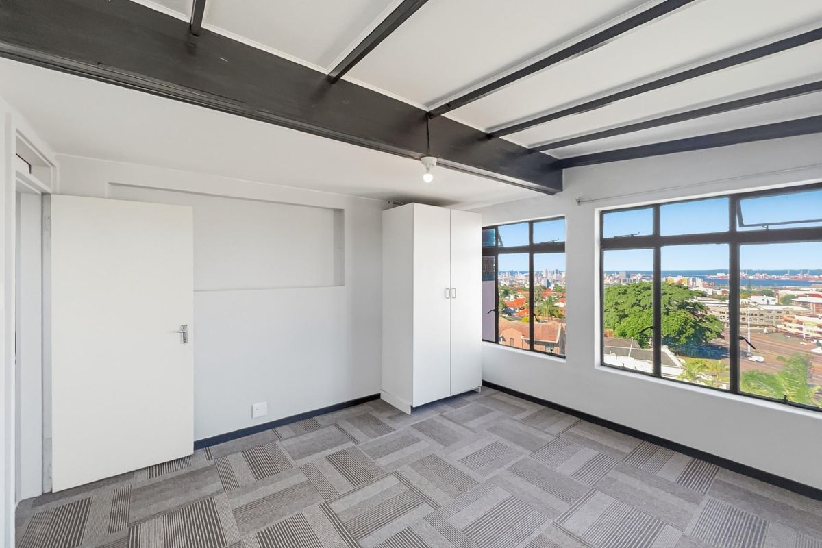 A bright bedroom featuring exposed ceiling beams, carpeted flooring, and large windows offering expansive city and coastal views.
