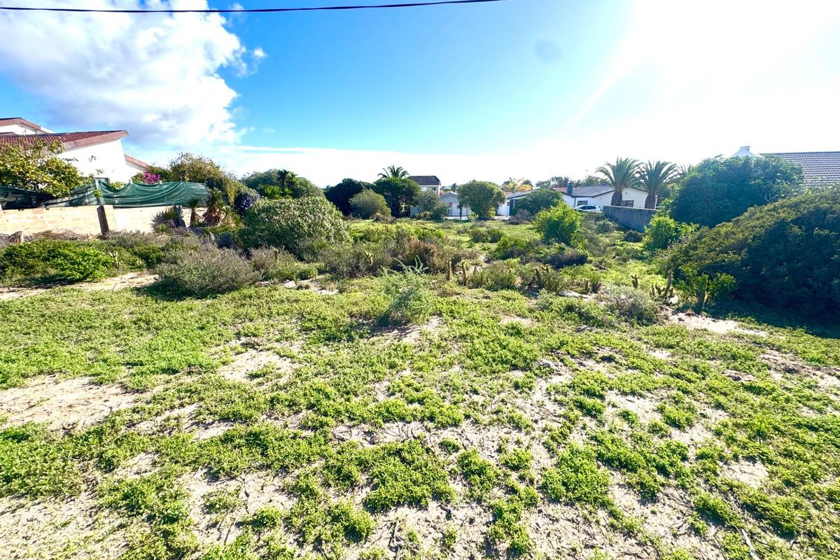 A vacant residential plot featuring natural vegetation and sandy ground, situated in a developed neighborhood setting.