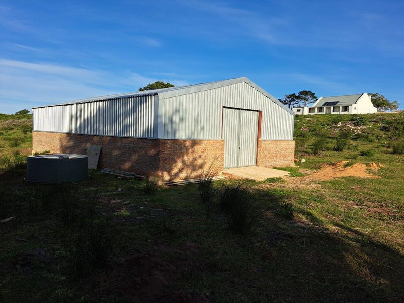 A functional farm outbuilding featuring a brick base and corrugated metal cladding, situated on a rural property with a main residence in the background.