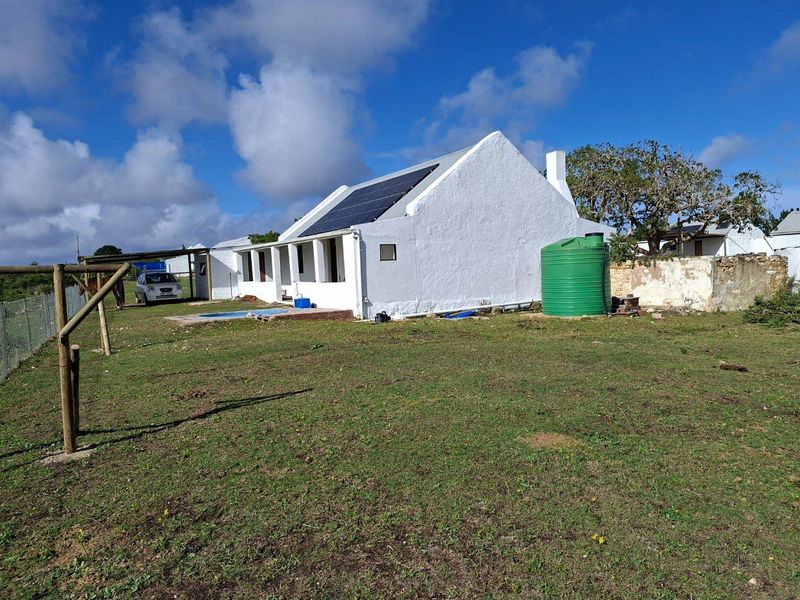 Rear view of a white farmhouse featuring solar panels, a water storage tank, and a small swimming pool set within a large grassy yard.
