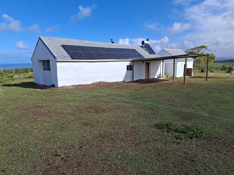 A rural farm cottage featuring a corrugated metal roof, integrated solar panels, a solar water heater, and an attached wooden pergola structure.