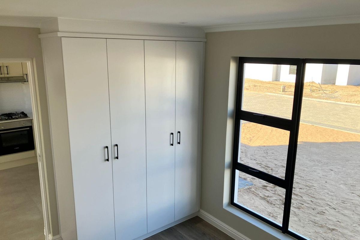 A bright bedroom featuring modern built-in cupboards with sleek handles, large black-framed windows, and durable wood-look flooring.