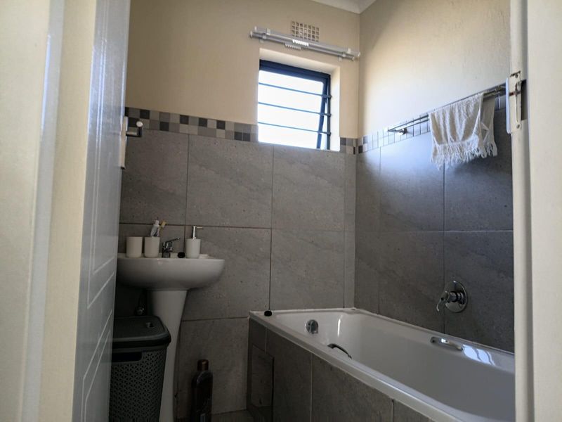 A functional bathroom featuring a pedestal basin, a built-in bathtub, and neutral grey wall tiling with a louver window for ventilation.