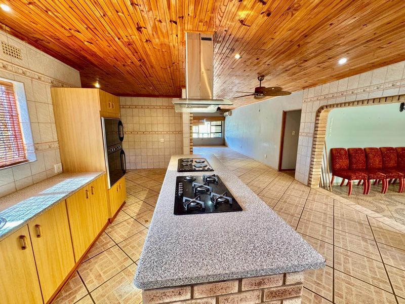 Spacious open-plan kitchen featuring a central island with built-in gas hob, stainless steel extractor fan, and integrated wall oven, set under a timber-clad ceiling.