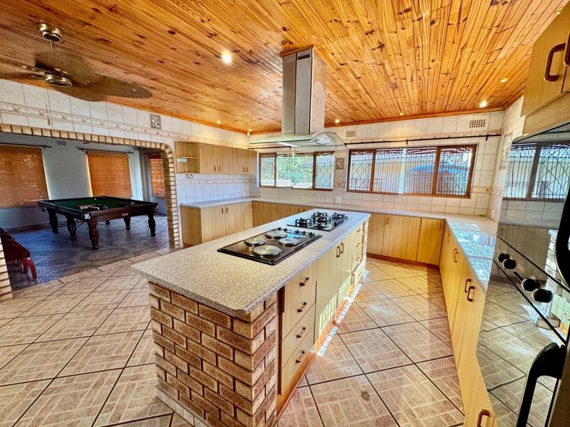 Spacious open-plan kitchen featuring a central island with built-in hob, ample cabinetry, and a distinctive timber-clad ceiling, flowing into an adjacent entertainment area.