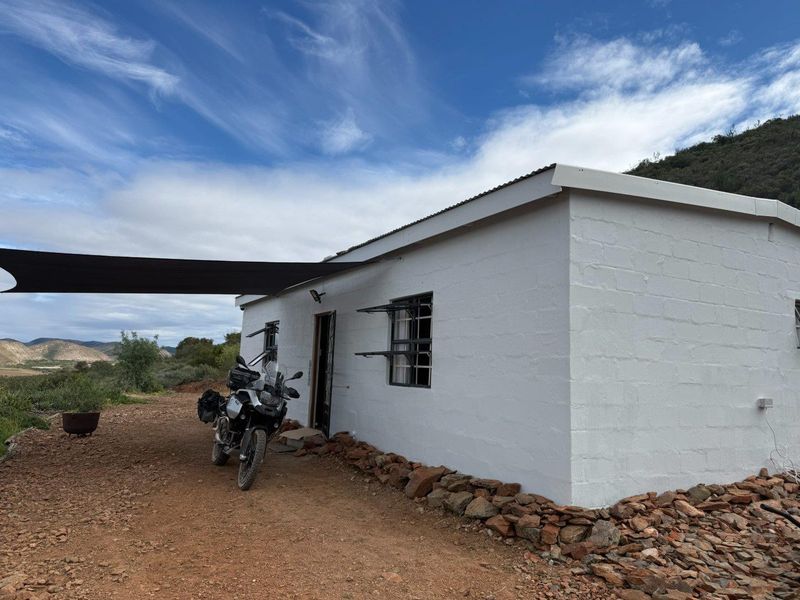 A simple white-painted farm cottage exterior featuring a shade sail, metal window frames, and a rural landscape setting.