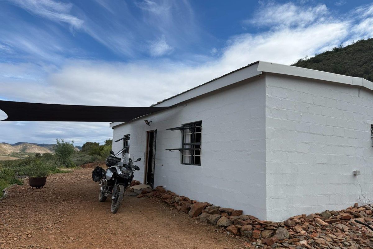A simple white-painted farm cottage exterior featuring a shade sail, metal window frames, and a rural landscape setting.