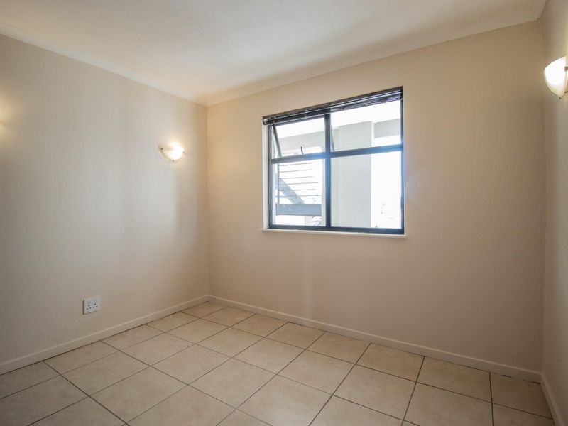 A clean, empty bedroom featuring neutral tiled flooring, wall-mounted lighting, and an aluminium-framed window providing natural light.