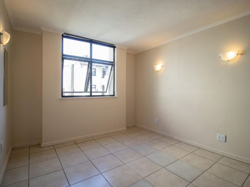 A bright, empty bedroom featuring neutral tiled flooring, wall-mounted lighting, and a large window providing natural light.