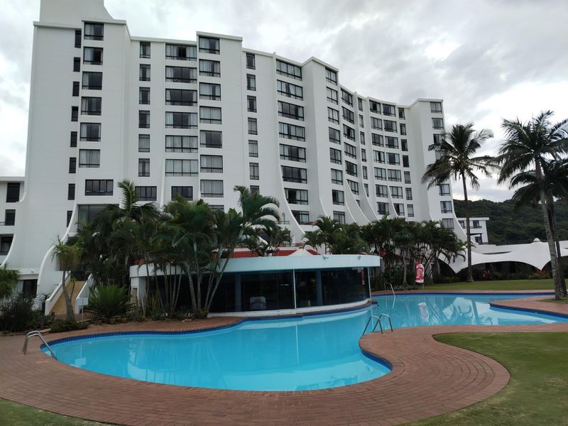 A view of the apartment building exterior featuring a large swimming pool, landscaped grounds, and palm trees.