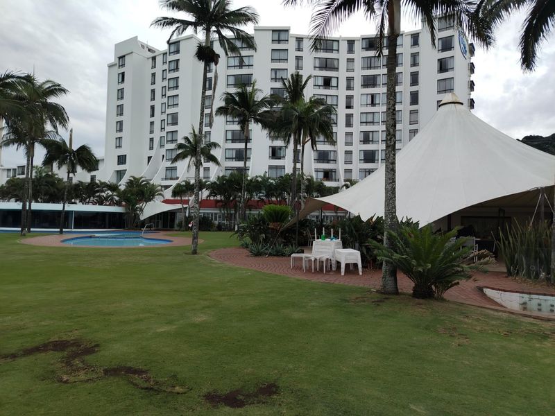 A view of the apartment complex exterior featuring a swimming pool, landscaped garden, and a large outdoor canopy structure.