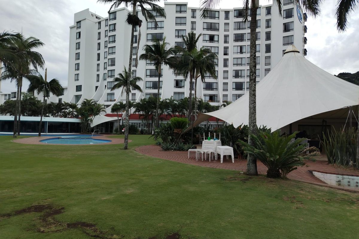A view of the apartment complex exterior featuring a swimming pool, landscaped garden, and a large outdoor canopy structure.