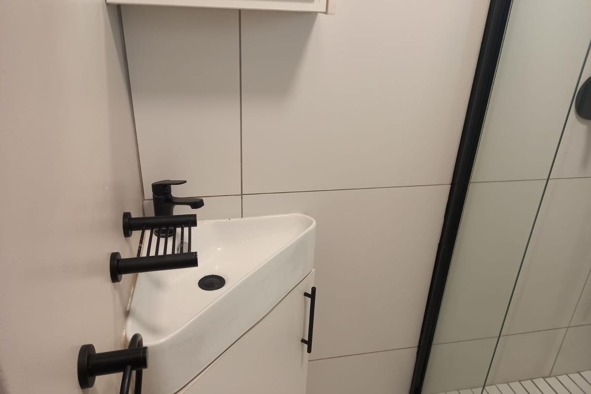 Compact bathroom featuring a corner vanity unit with a modern black faucet, wall-mounted accessories, and a glass-enclosed shower with tiled walls.