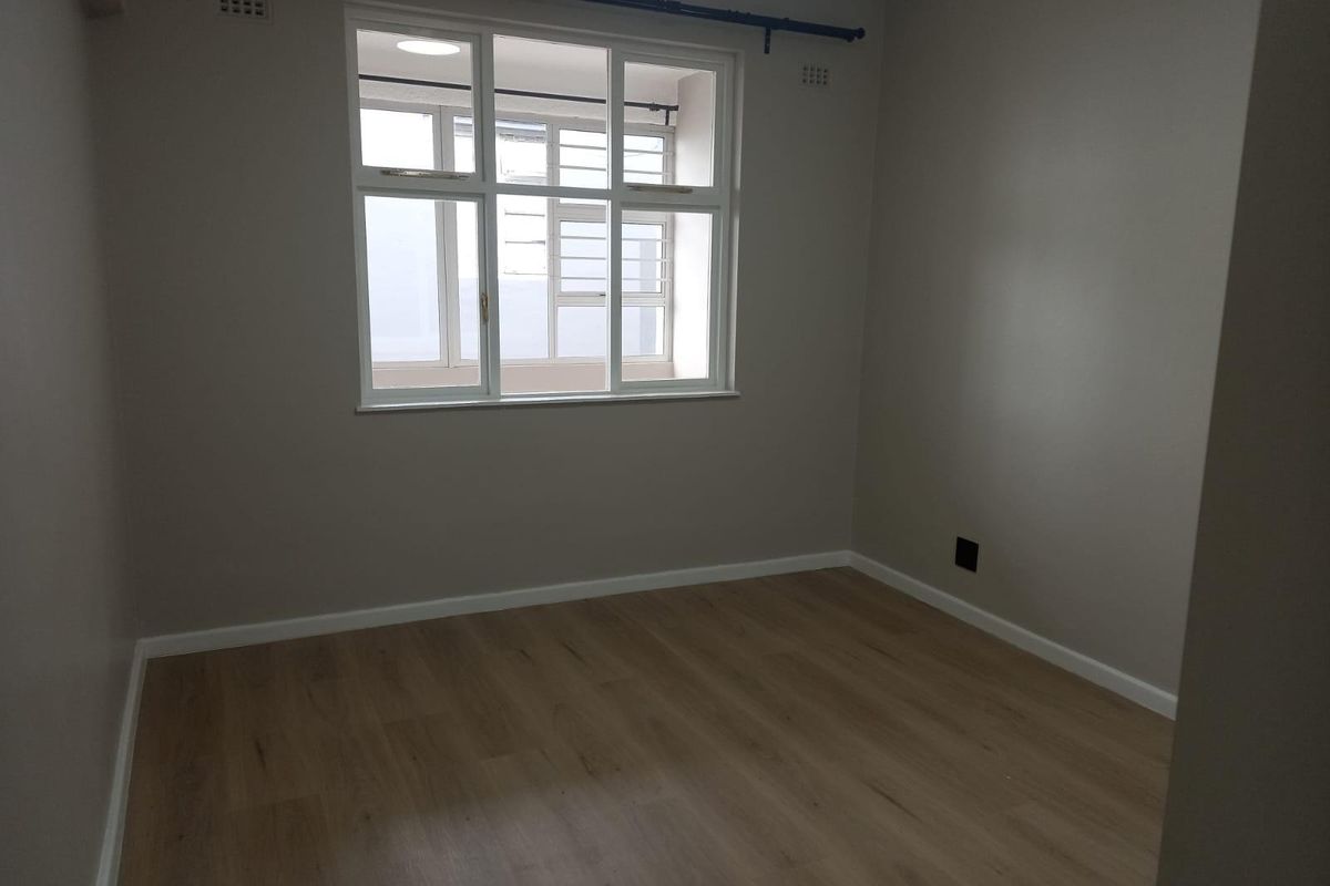 A clean, empty bedroom featuring neutral wall paint, modern wood-look laminate flooring, and a large window with security bars.
