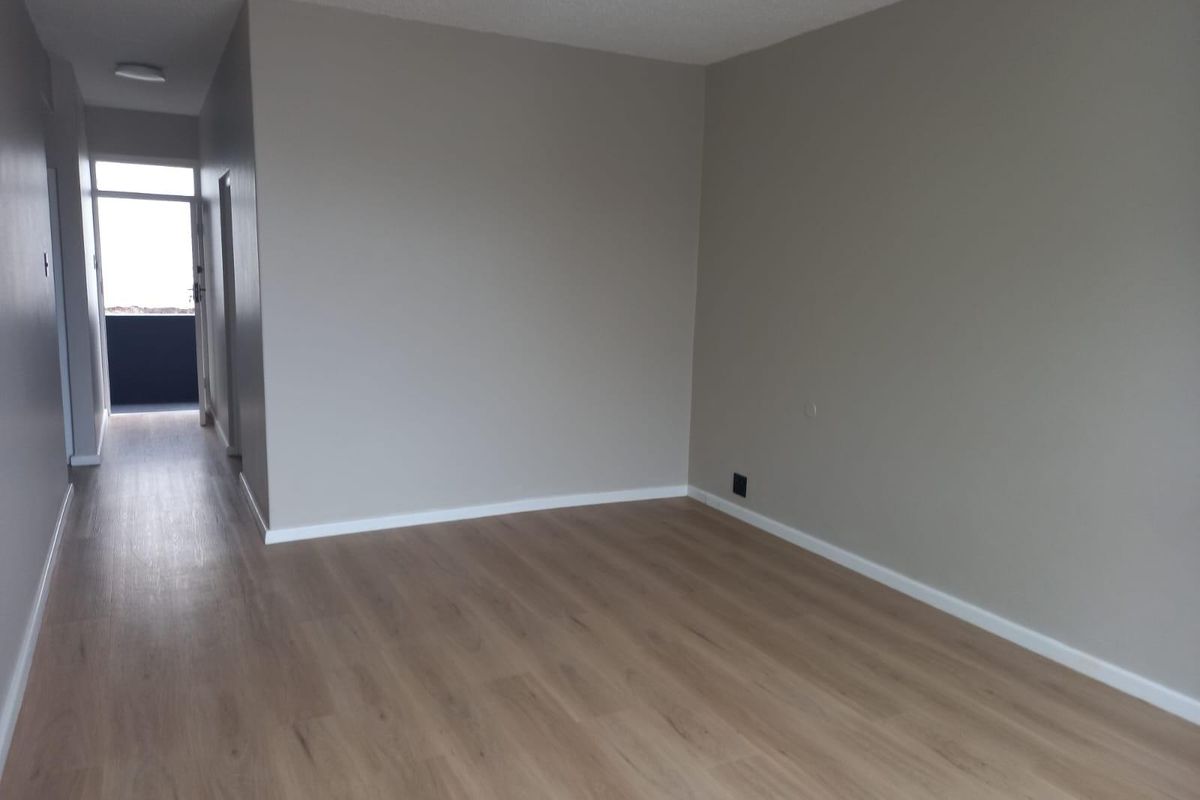 A bright, empty living area featuring modern wood-look laminate flooring and neutral wall tones, leading to a hallway with access to an exterior balcony.