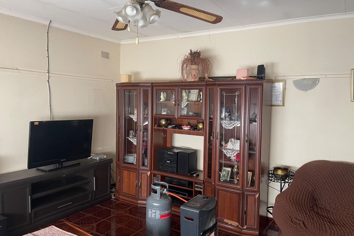 A living room featuring tiled flooring, a ceiling fan, and wall-mounted lighting, showing signs of wear and dated finishes.