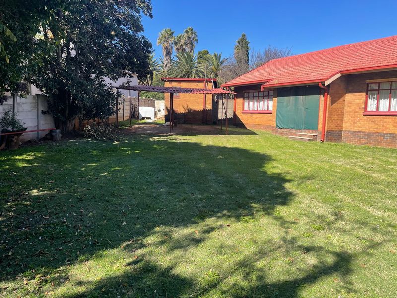 A spacious rear garden featuring a brick house with a red tiled roof, a metal pergola structure, and established trees.