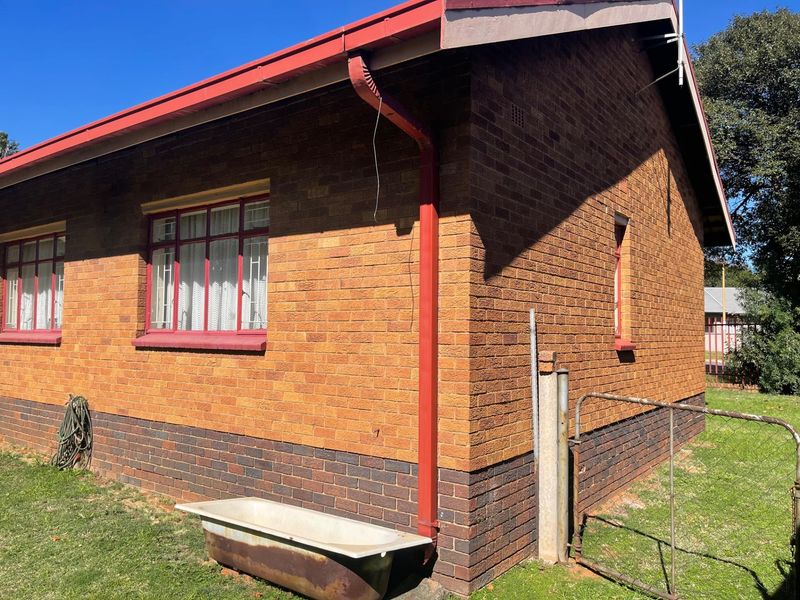 Exterior view of a brick house featuring red-painted window frames, a metal roof, and a side garden area.