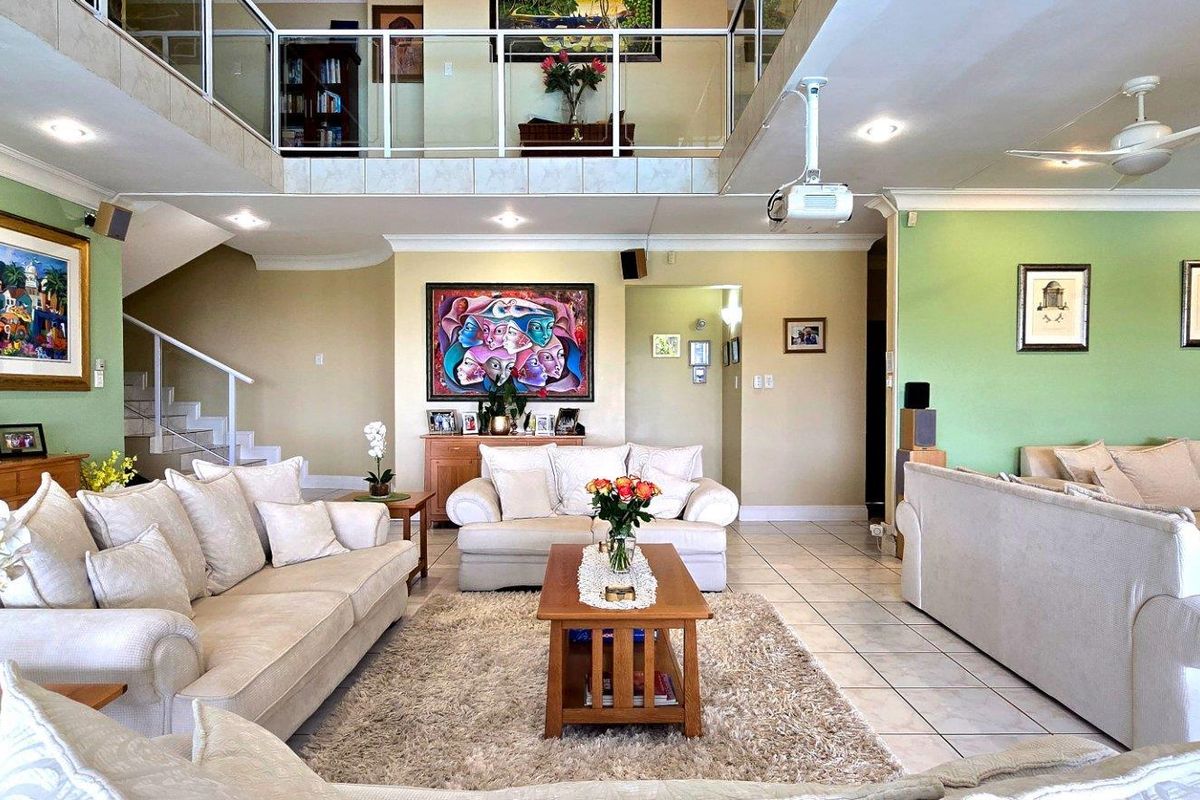 Spacious double-volume living area featuring tiled flooring, a mezzanine level with glass balustrade, and integrated ceiling fan and recessed lighting.