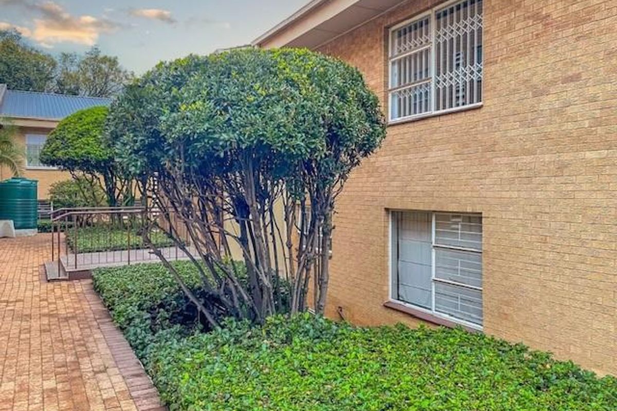 Exterior view of the apartment building featuring face brick facade, security bars on windows, and paved walkway alongside landscaped garden beds.