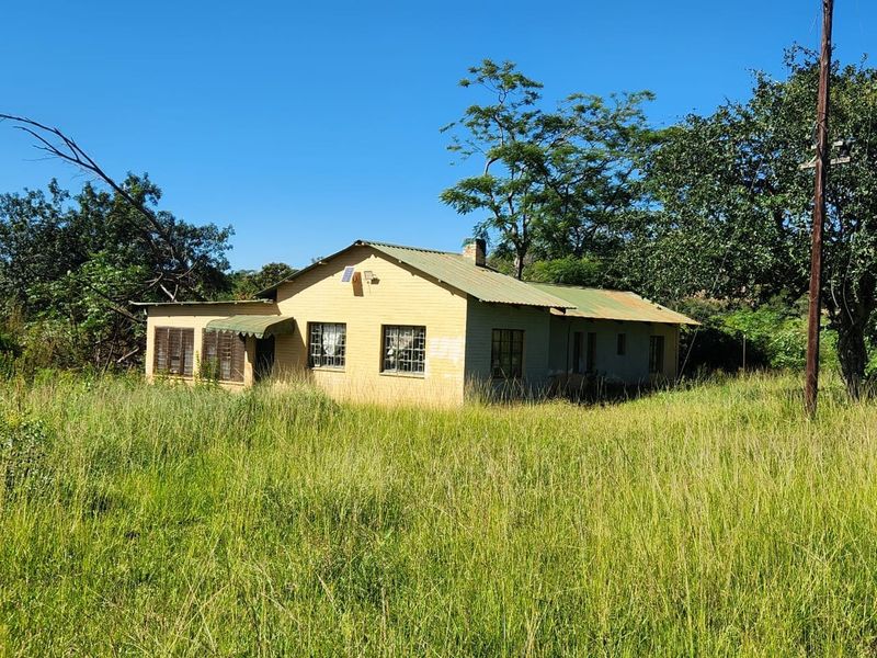 A detached farm cottage featuring a corrugated metal roof, chimney, and security bars, set within an expansive, overgrown rural landscape.