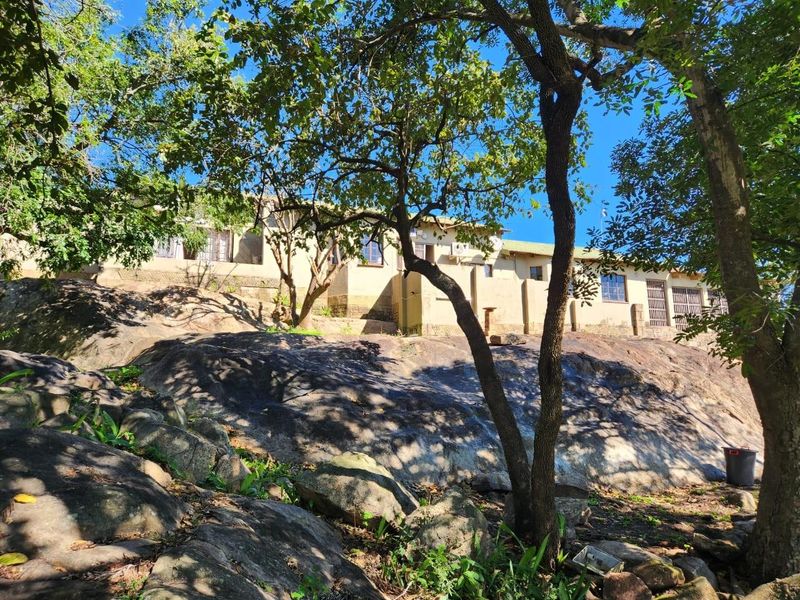 A view of the rear exterior of the farm dwelling, featuring a natural rocky landscape and mature trees surrounding the building structure.