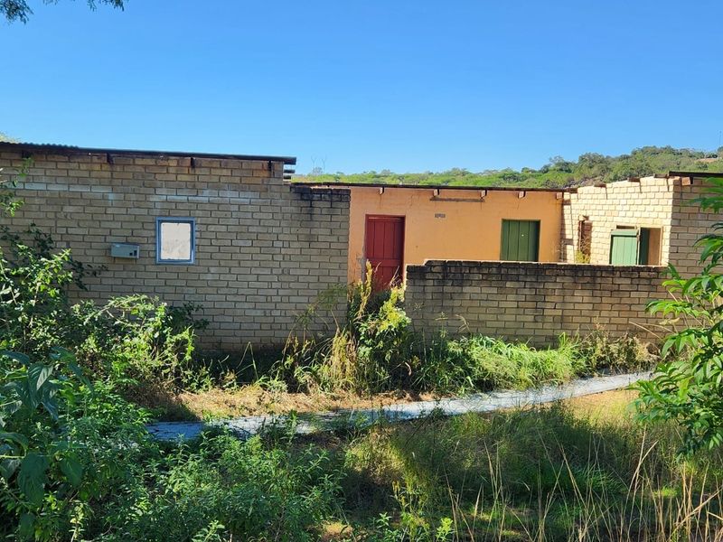 Rear view of a farm building featuring exposed brick walls, metal roofing, and multiple access doors, set within an overgrown garden area.