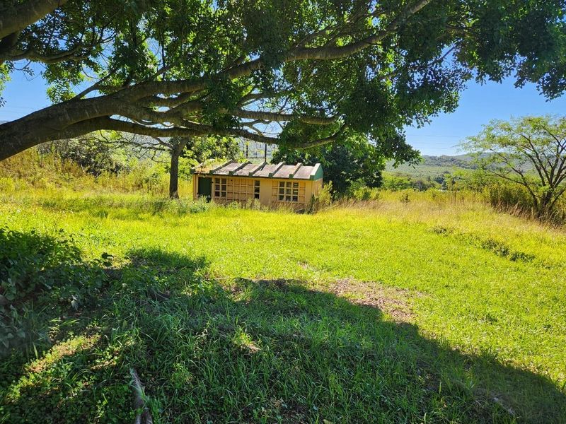 A small, weathered outbuilding situated on a rural farm property, surrounded by open grassy fields and mature trees.