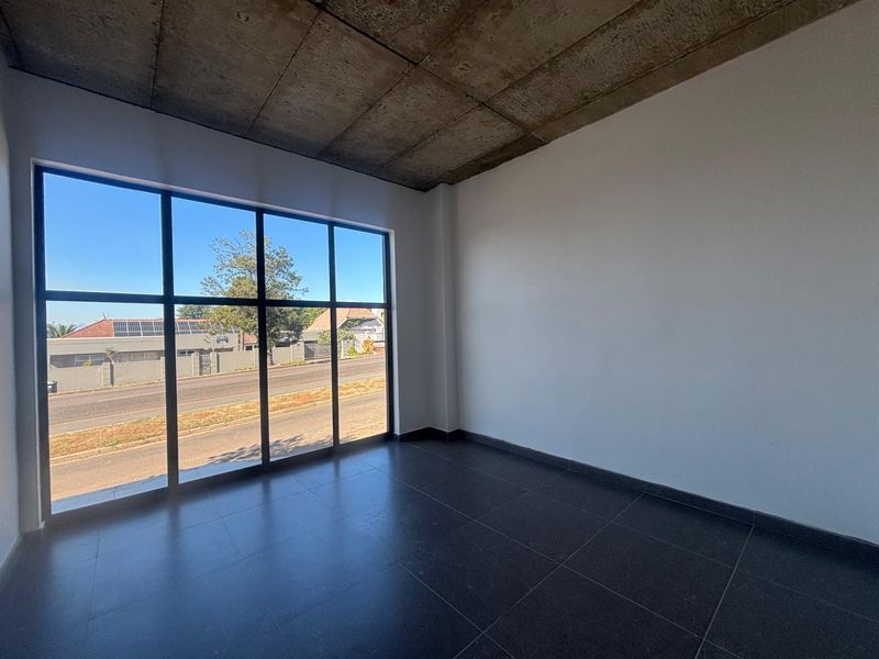A bright, modern office space featuring an exposed concrete ceiling, large floor-to-ceiling windows with street views, and dark tiled flooring.