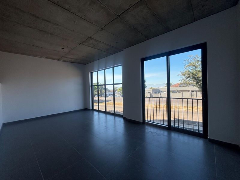 Modern open-plan office space featuring an exposed concrete ceiling, large windows for natural light, and sleek tiled flooring with access to a small balcony.