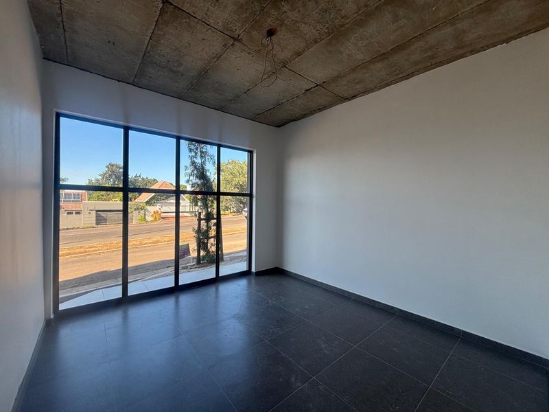 A bright, modern office space featuring large floor-to-ceiling windows, dark tiled flooring, and an industrial-style exposed concrete ceiling.