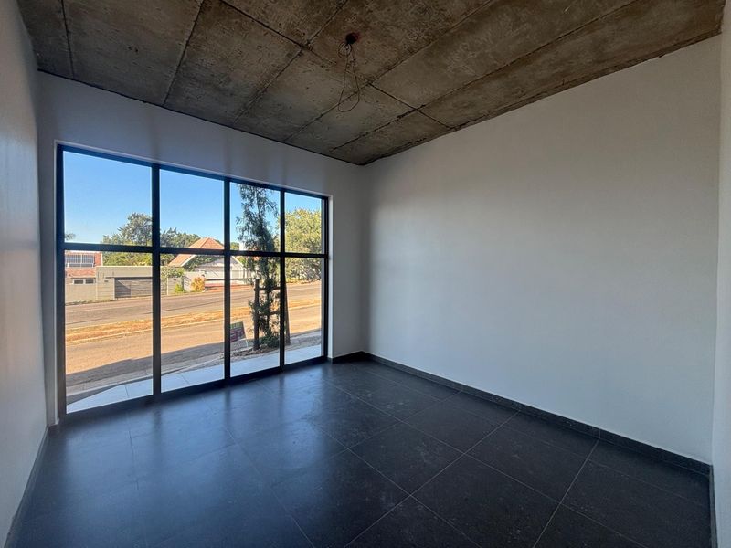 A bright, empty office space featuring large floor-to-ceiling windows, dark tiled flooring, and an industrial-style exposed concrete ceiling.
