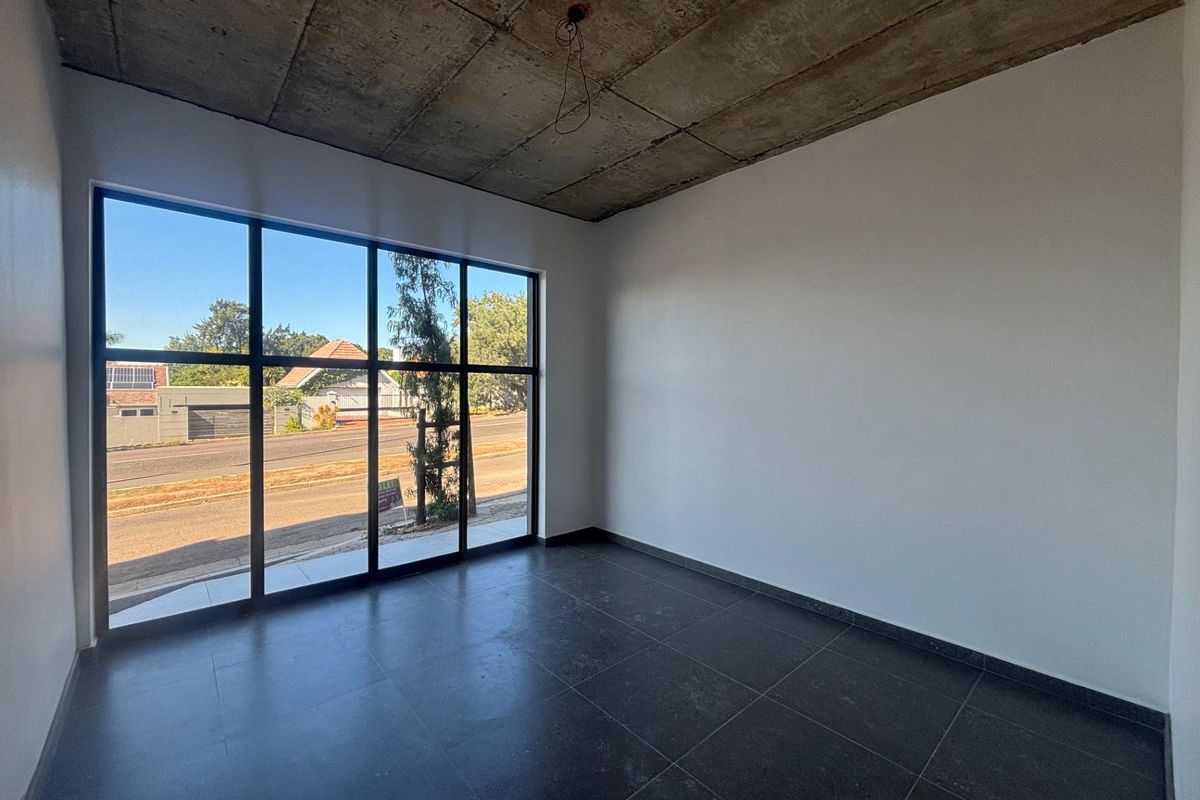 A bright, empty office space featuring large floor-to-ceiling windows, dark tiled flooring, and an industrial-style exposed concrete ceiling.