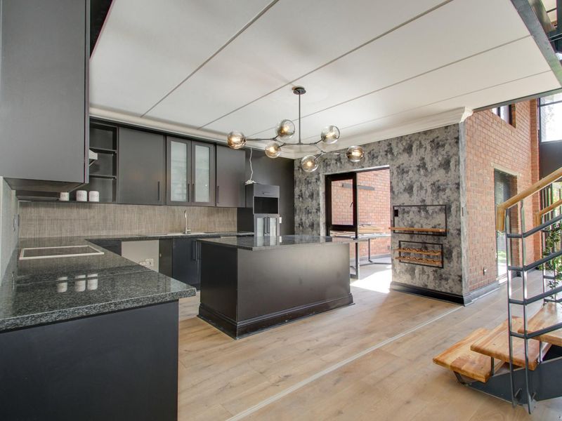 Modern kitchen featuring dark cabinetry, granite countertops, a central island, and exposed brick accents, seamlessly integrated with an open-plan living area and staircase.