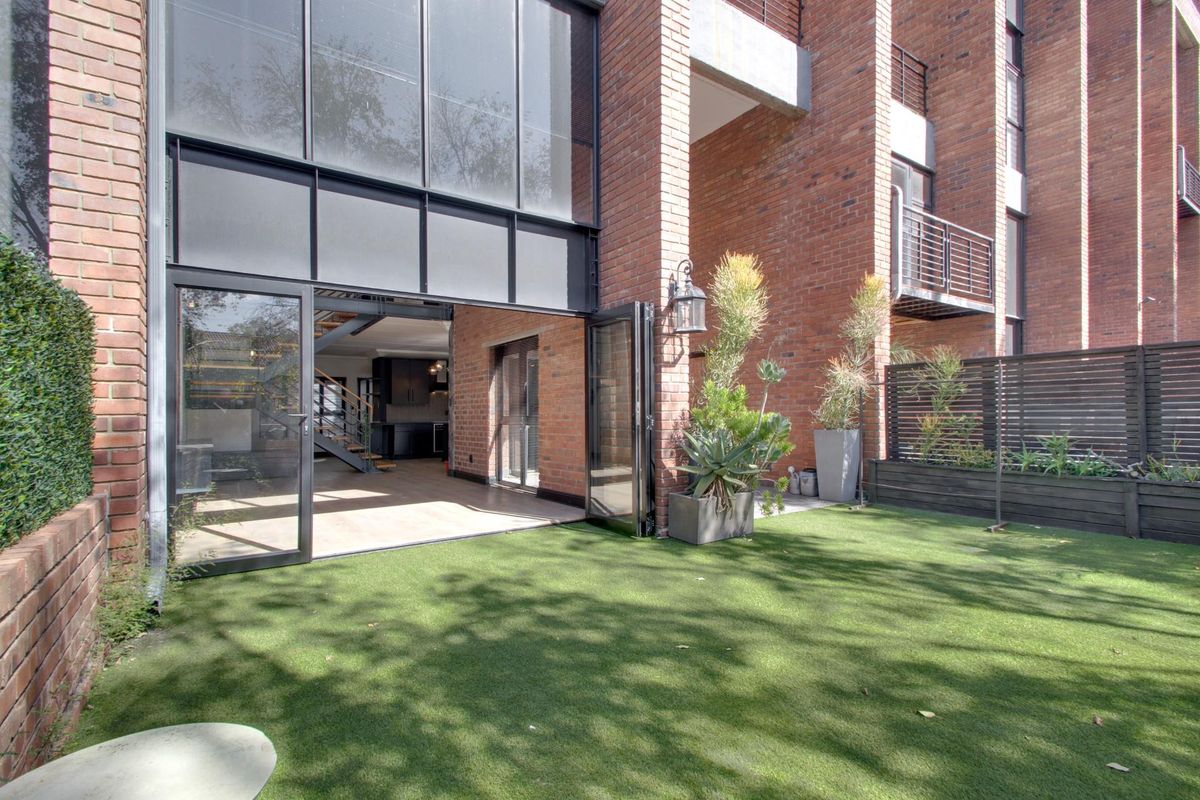 A modern rear exterior view featuring exposed brickwork, large bi-fold glass doors opening to an artificial turf garden, and a contemporary steel staircase.