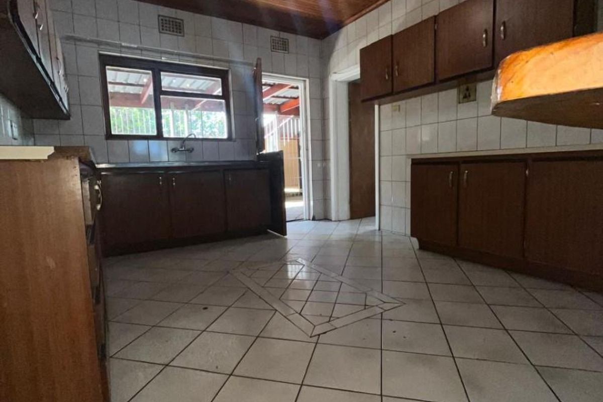 A dated kitchen featuring wooden cabinetry, tiled flooring with decorative inlay, and a wooden panelled ceiling, with direct access to an outdoor area.