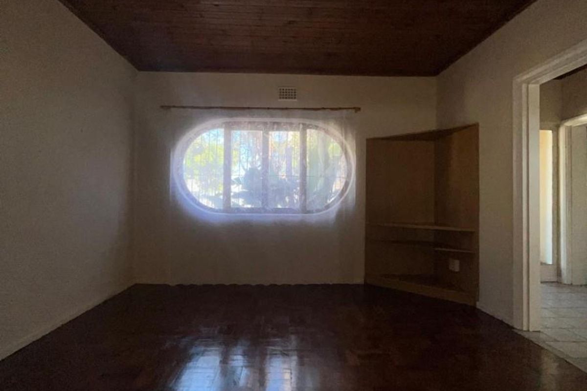 A spacious living room featuring polished parquet flooring, a distinctive oval window, and a timber-paneled ceiling with built-in corner shelving.