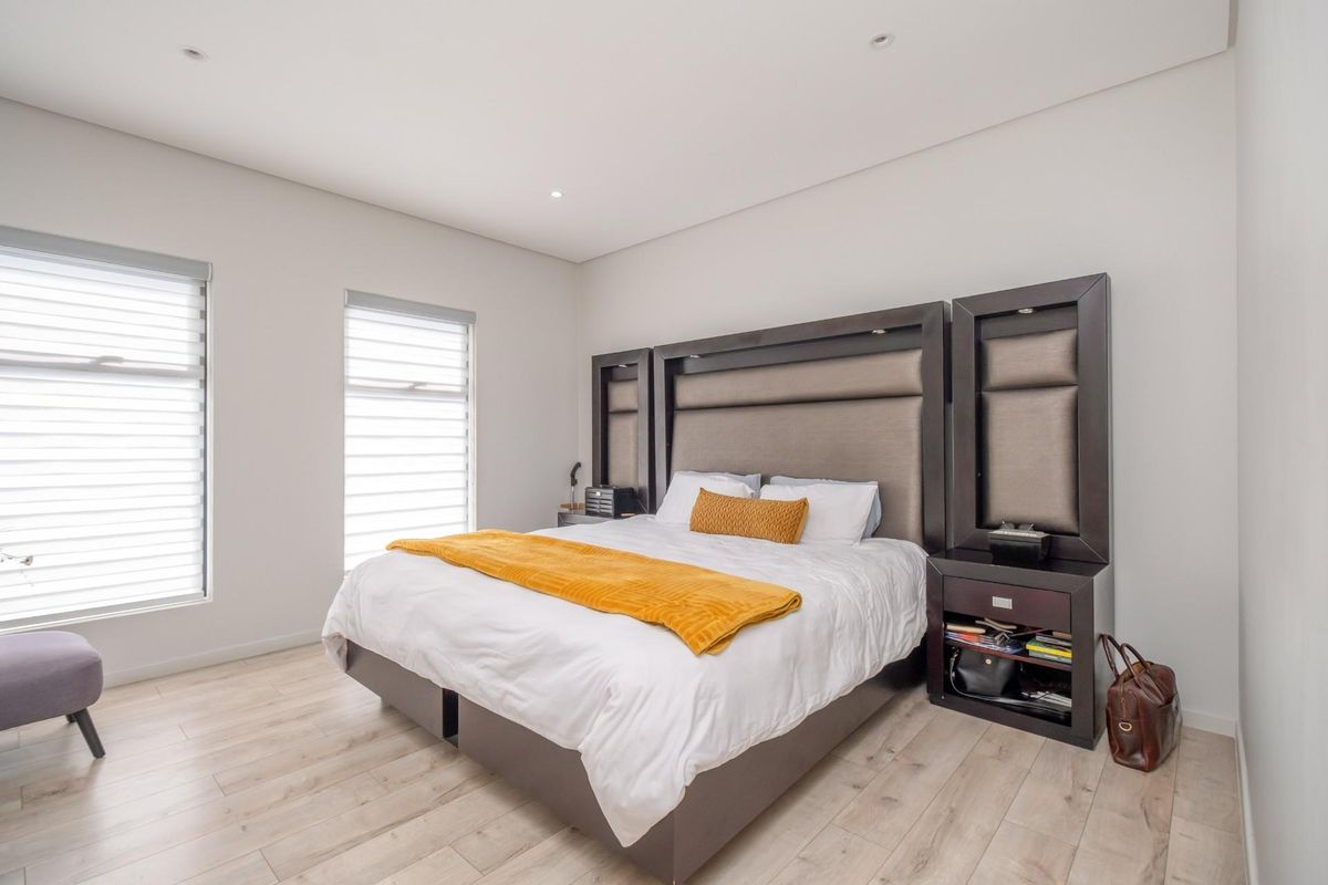 A modern, well-lit bedroom featuring light wood-look flooring, recessed ceiling lighting, and fitted window blinds.