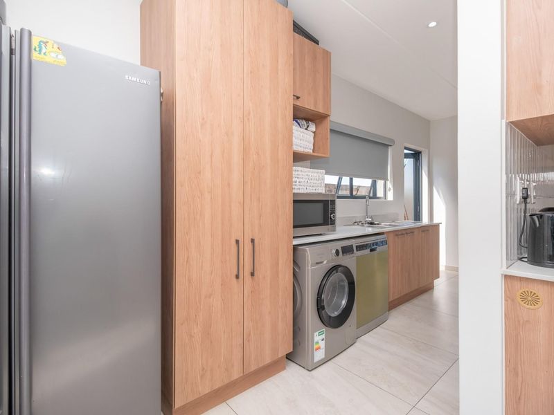A functional scullery area featuring light wood cabinetry, integrated appliance spaces, a stainless steel sink, and tiled flooring.