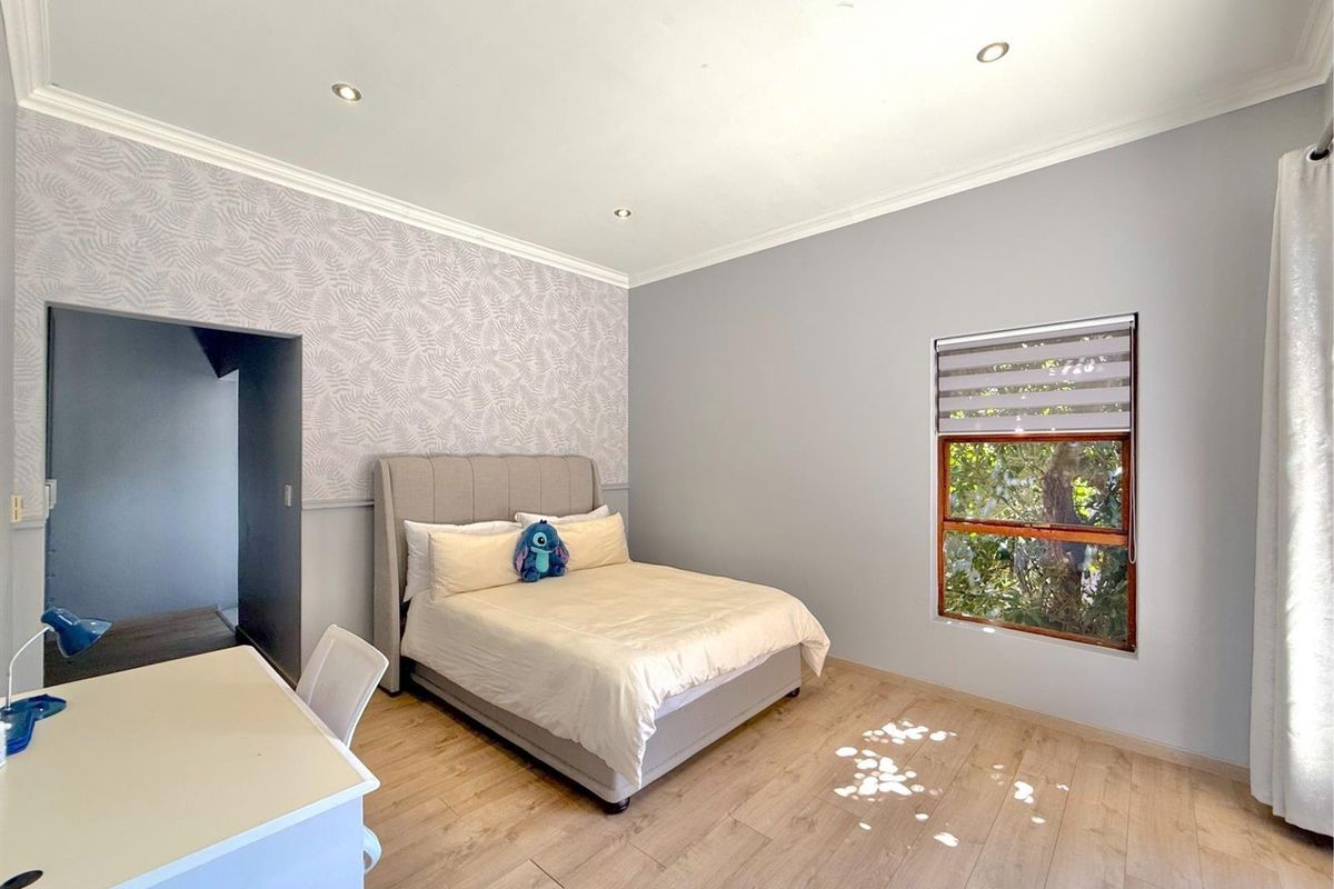 Spacious bedroom featuring light wood-look flooring, recessed ceiling lighting, and a feature wallpaper wall with a window overlooking greenery.