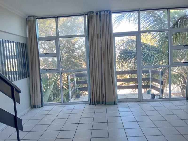 Bright living area featuring large floor-to-ceiling windows and glass doors leading to a private balcony, with neutral tiled flooring throughout.